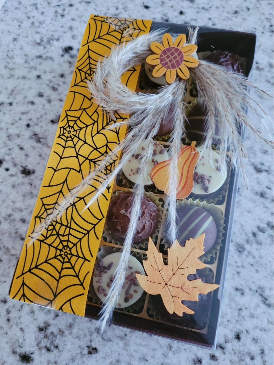 Boîte de pralinés assortis avec un ruban jaune à toiles d'araignée, décorée d'herbes séchées, d'un tournesol, d'une courge et d'une feuille d'érable en bois